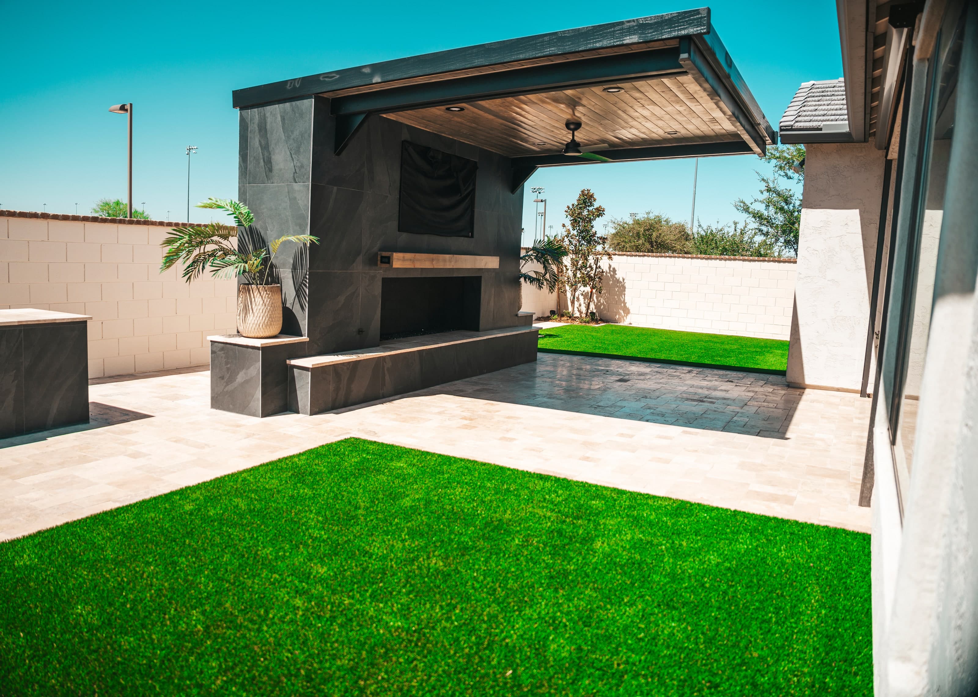Built-in shade structure with a fireplace wall over a travertine patio, beside a turf lawn in a Gilbert backyard