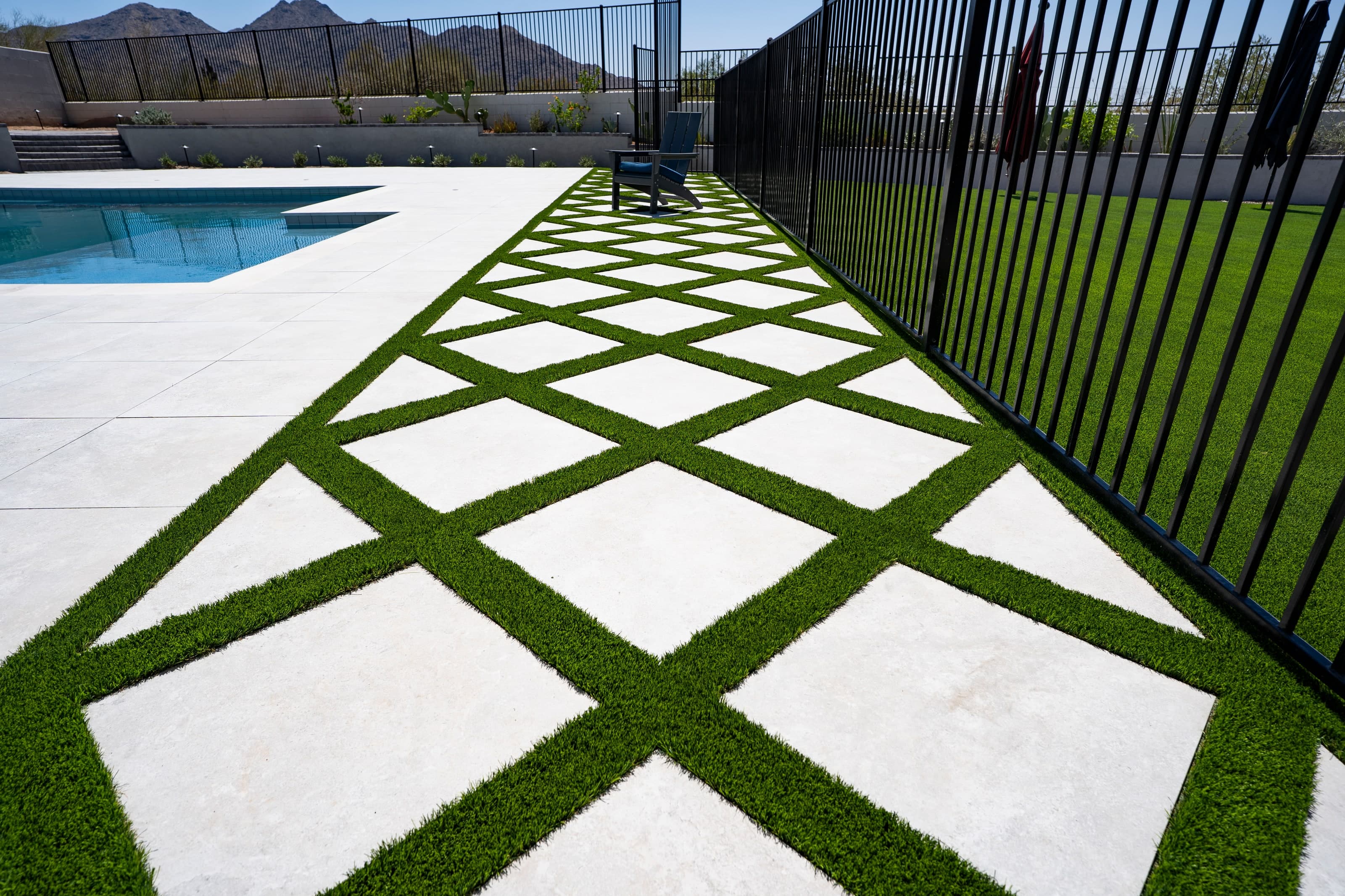 A lattice-pattern turf-and-paver strip running beside a black metal pool fence, with the pool deck on the left and the lawn behind the fence