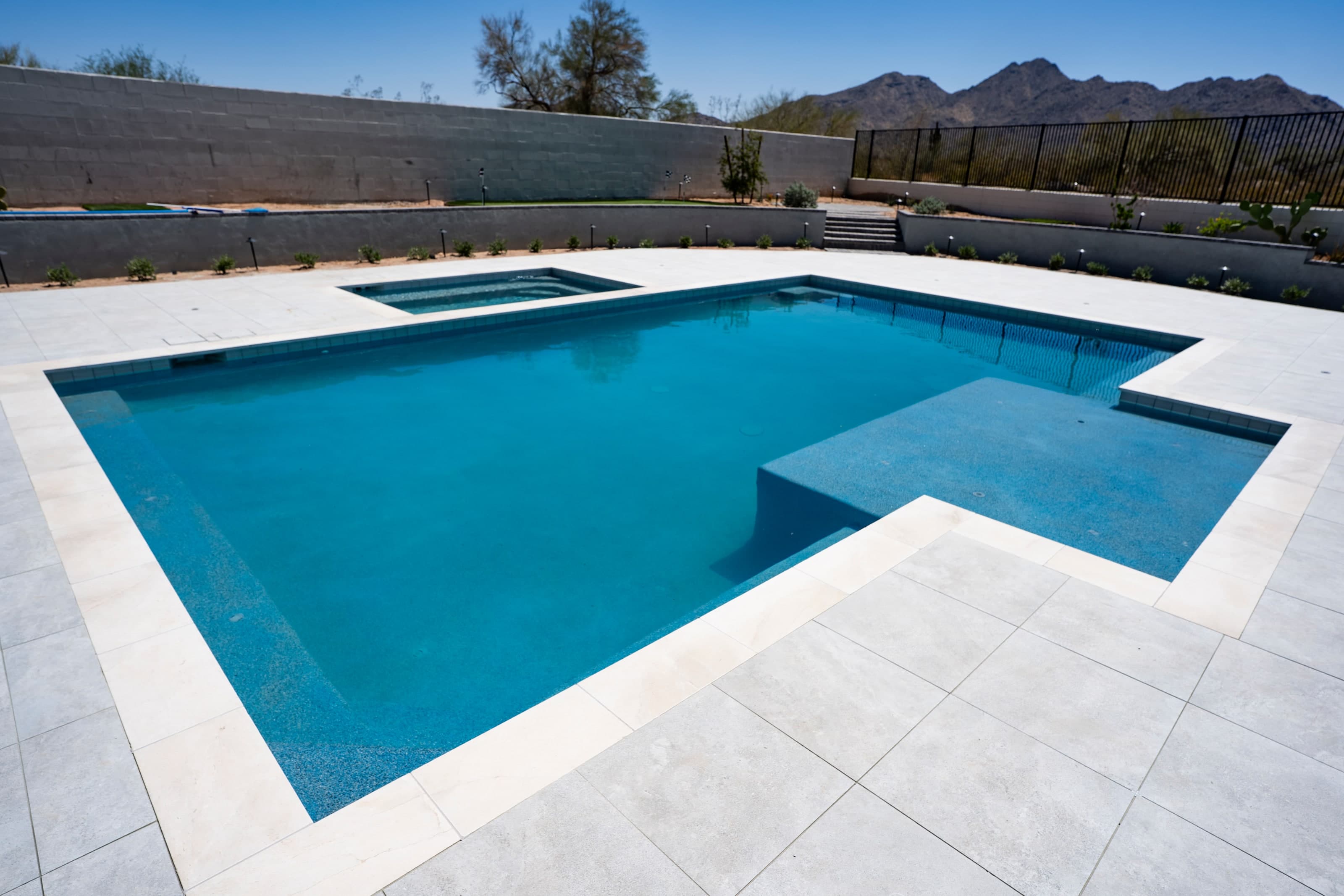 A pool and raised-edge spa surrounded by wide, light-colored large-format decking, with a block wall and mountains behind