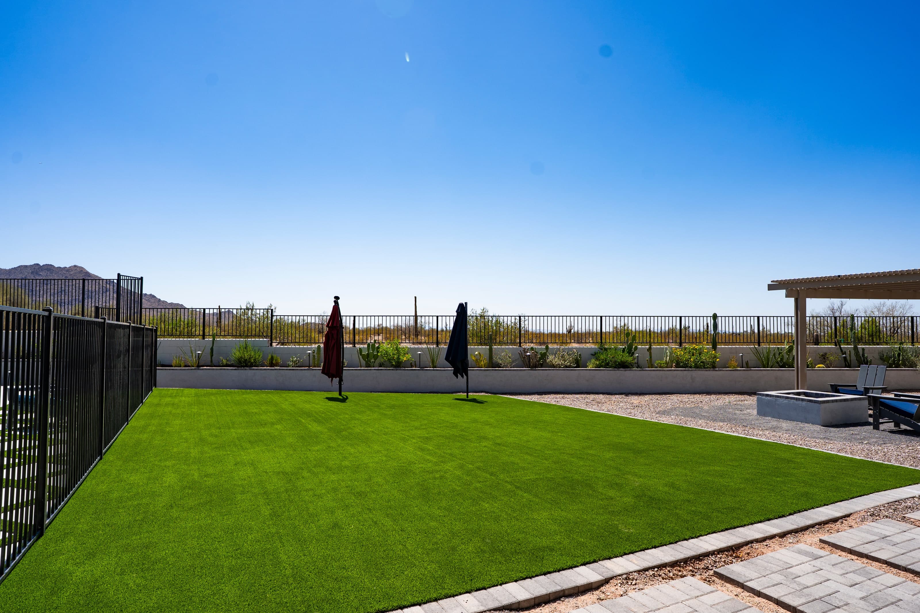 An open turf lawn with a pool fence on the left, two closed umbrellas, and a patio cover and desert planting along the back