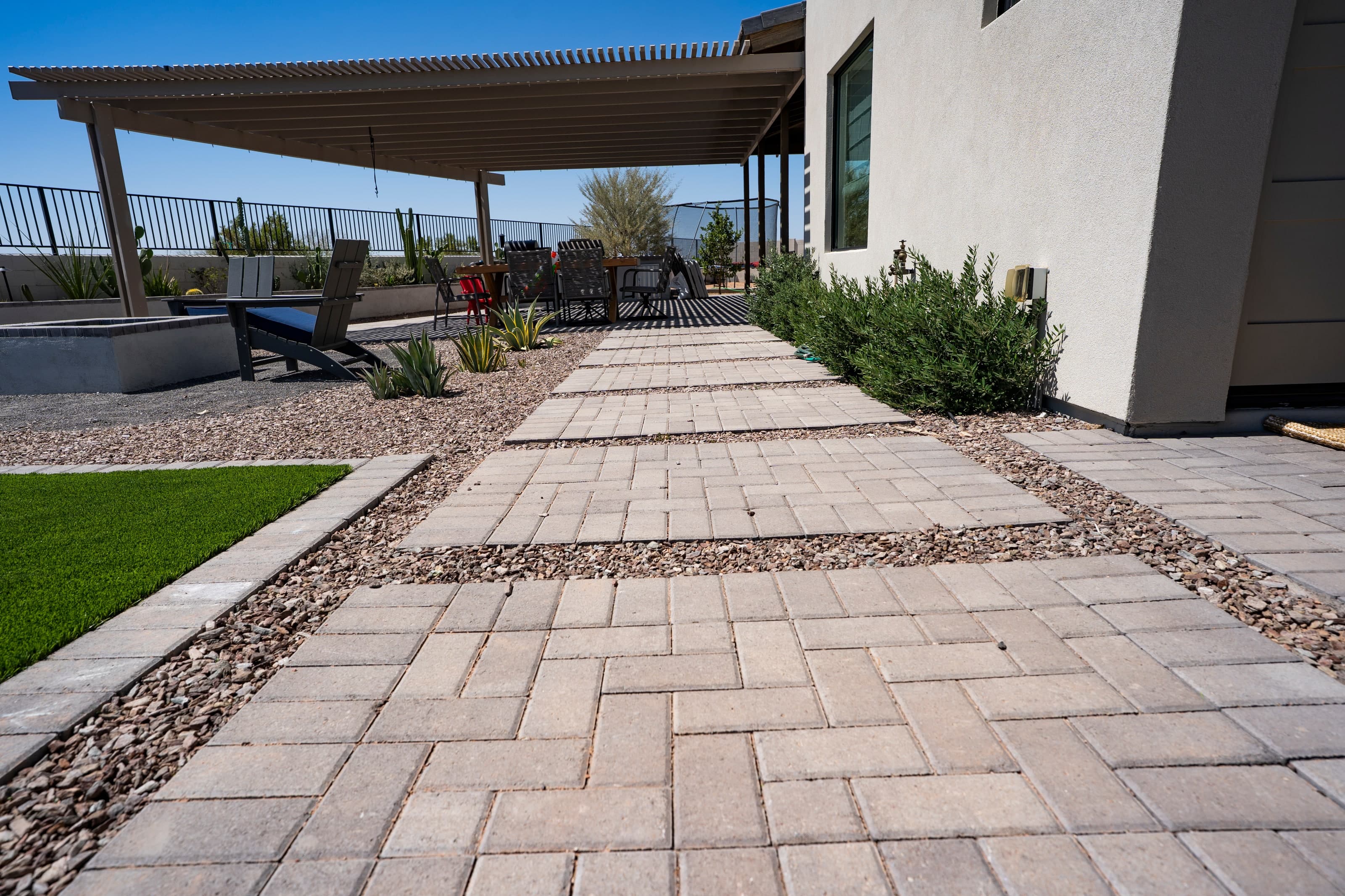 A paver walkway running along the back of the house beside the lawn, with gravel and planting on either side and a patio cover overhead