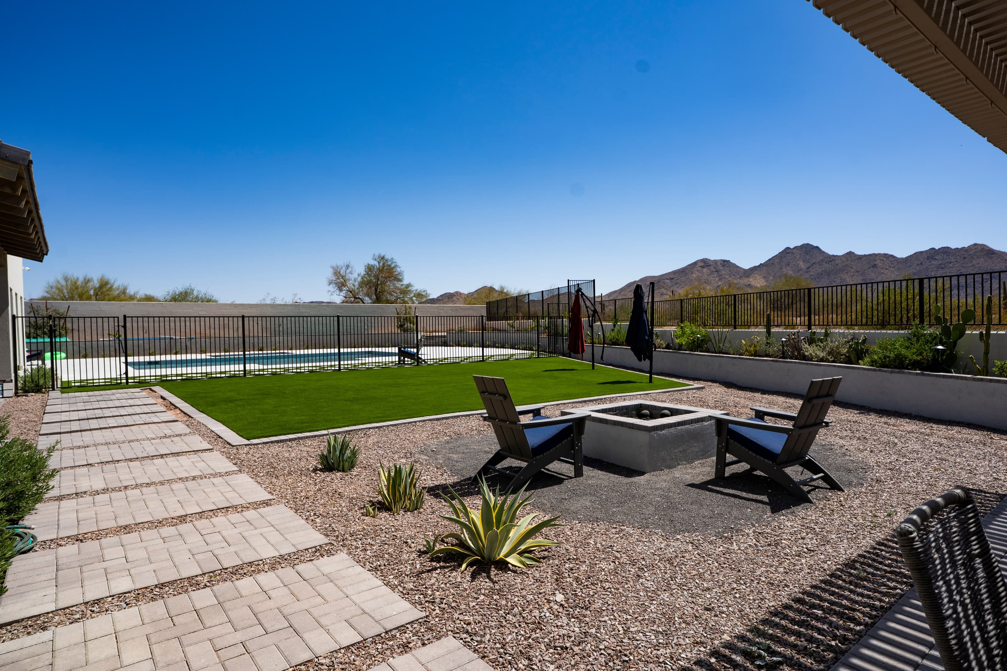 Paver stepping path crossing gravel to a small square fire pit with two chairs, beside the turf lawn, with the pool fence beyond
