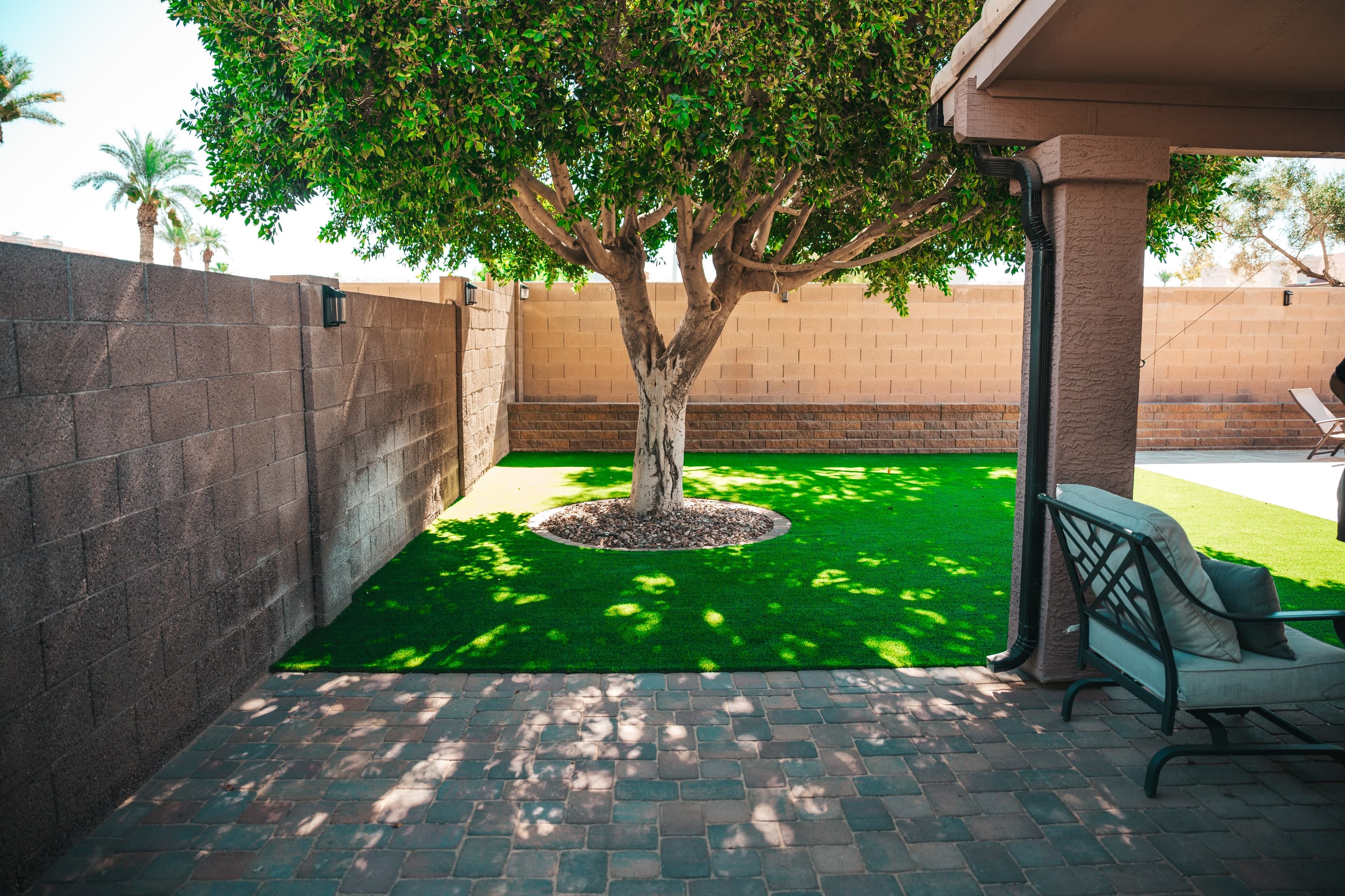 A paver patio beside a covered porch, looking onto a shaded turf lawn under a large tree