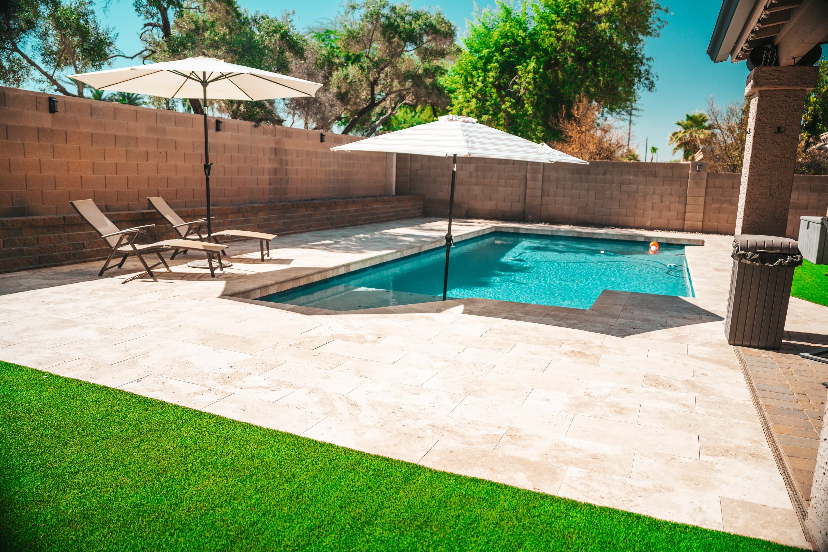 Travertine pool deck with lounge chairs and umbrellas beside turf in a Chandler backyard