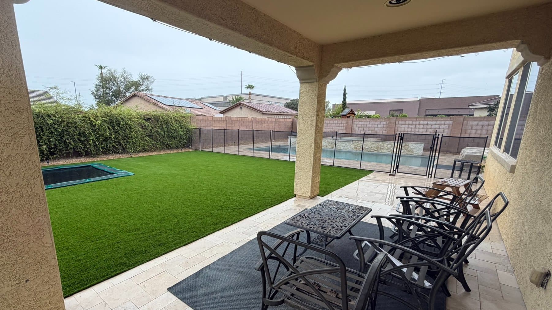 View from a covered travertine patio with outdoor chairs, across a turf lawn and sunken trampoline to a pool behind a safety fence
