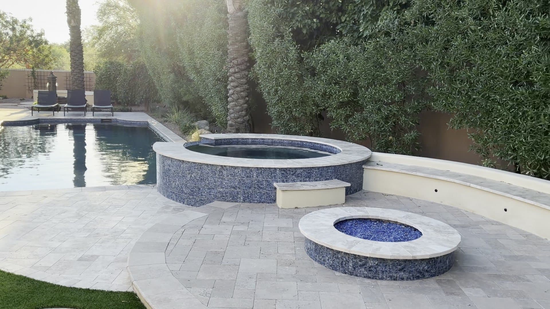 A round fire pit with blue glass on a travertine deck, in front of a raised spa and a long curved bench, with the pool behind