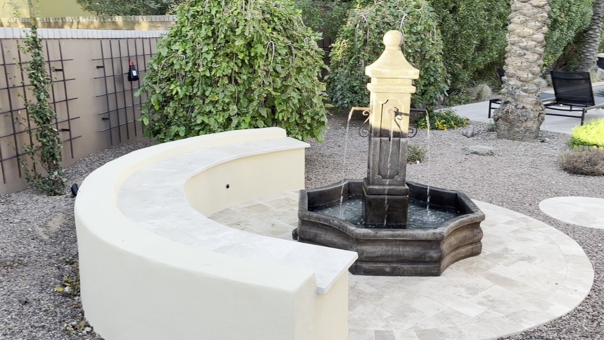 A tiered stone fountain beside a curved cream bench on a travertine pad, with a round stepping pad and gravel planting around it