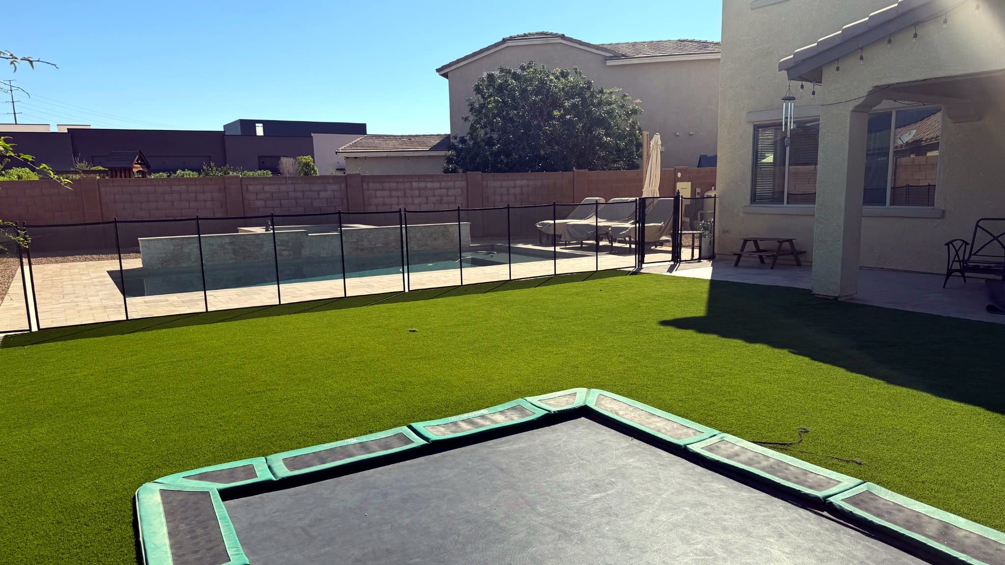 Sunny backyard with a turf lawn and the edge of a sunken trampoline in the foreground, a pool behind a mesh safety fence, and a covered patio with seating along the house
