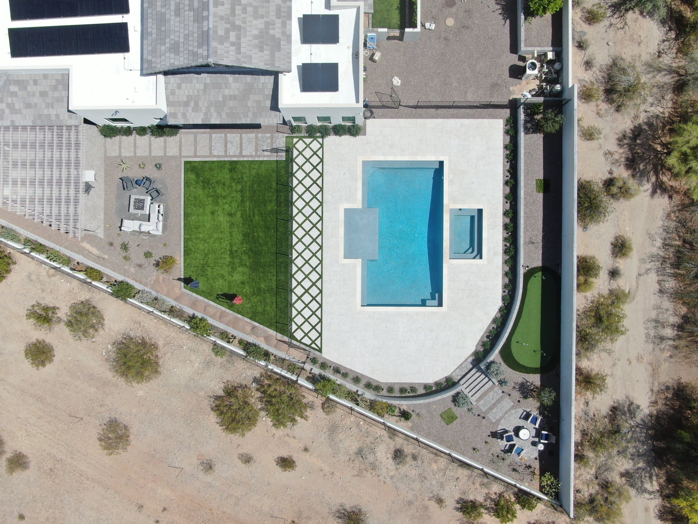 Straight-down drone view of the backyard: the house at the top, a paver path along the back of the house, a small fire-pit lounge and lawn on the left, a lattice-pattern strip and fence between the lawn and the pool deck, the pool and spa in the middle, and a putting green and round fire-pit patio along the right-hand wall