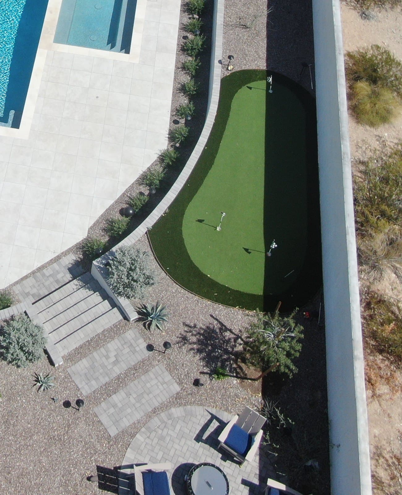 Overhead view of a putting green in a strip of side yard between a curved wall at the edge of a pool deck and a tall perimeter wall, with paver steps and steppers leading to a round fire pit on a circular paver patio