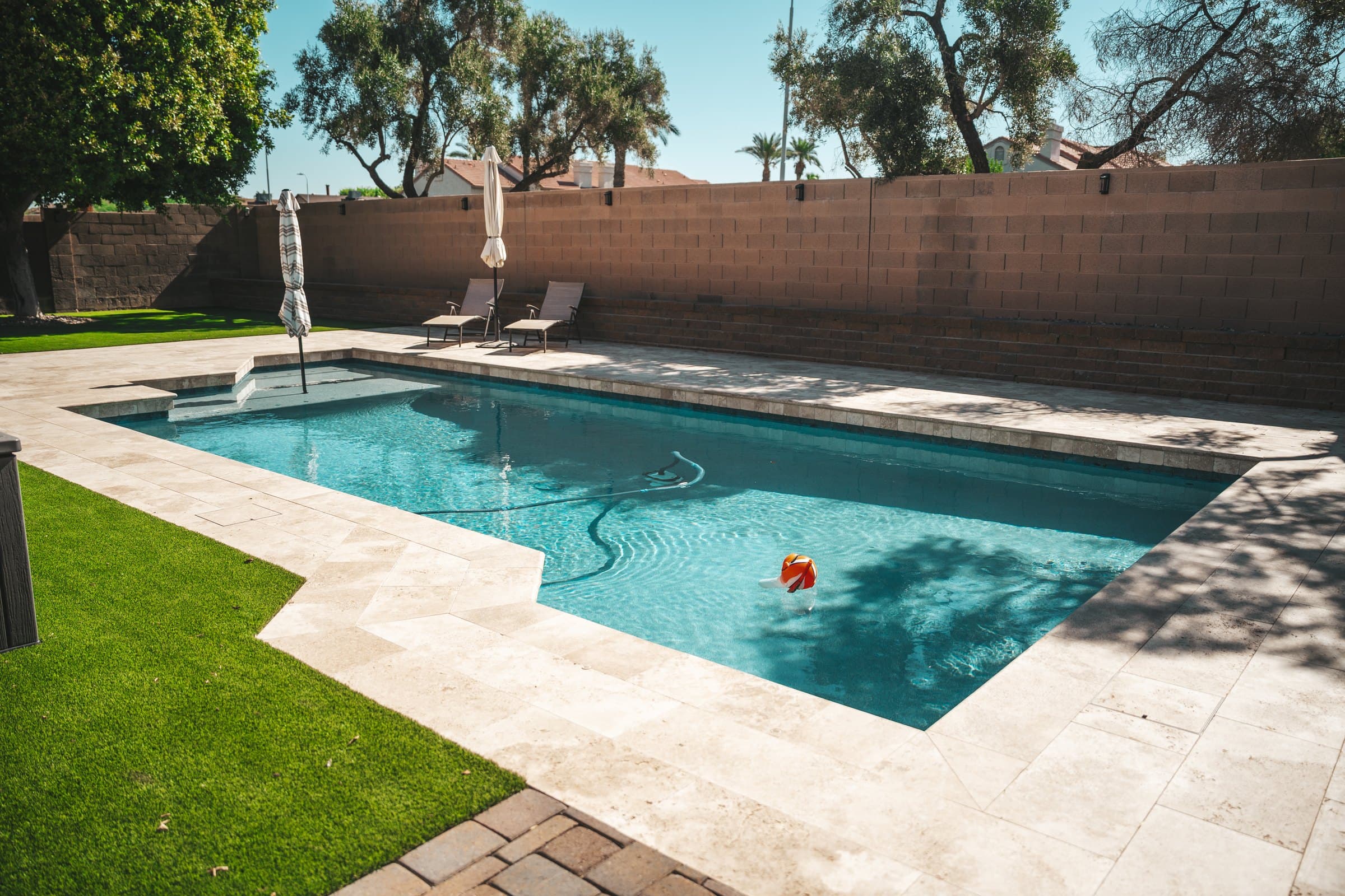 Light travertine decking around a rectangular pool, with turf following the deck's stepped edge and brown pavers in the foreground