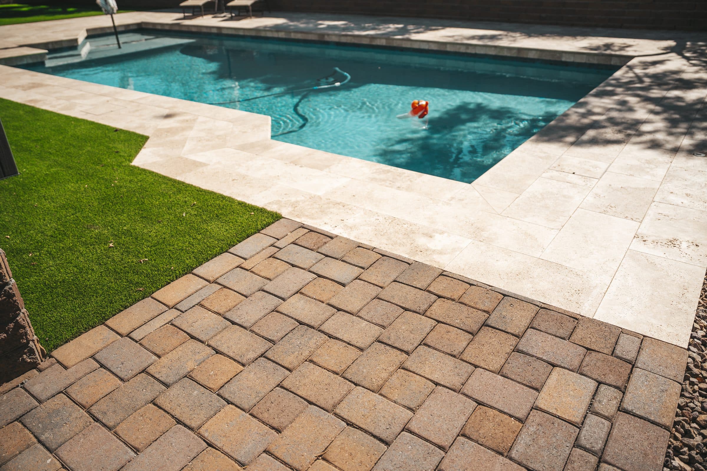 Brown concrete pavers meeting a light travertine pool deck, with turf along one side
